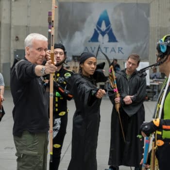 Behind-the-scenes photo from 'Avatar: Fire and Ash' featuring director James Cameron instructing actor Zoe Saldana and other cast members, all in motion capture suits, set in a studio environment.