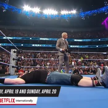 John Cena is laid out on the wrestling mat in the ring during a WWE SmackDown event, while Cody Rhodes stands nearby in a suit, addressing the crowd. The arena is filled with spectators and bright lights overhead.