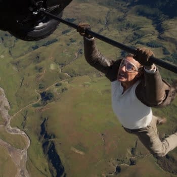 A scene from 'Mission: Impossible - The Final Reckoning' featuring Tom Cruise as Ethan Hunt, dramatically hanging from a helicopter above a vast green landscape.