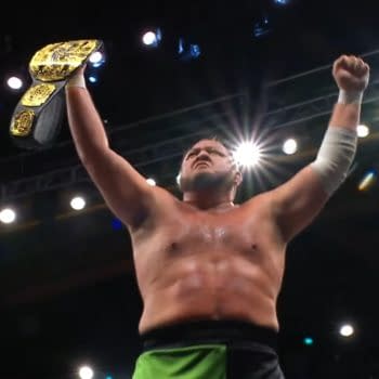 Samoa Joe stands with a championship belt raised high in his right hand, celebrating a victory at the end of AEW Dynamite, with bright arena lights shining around him.