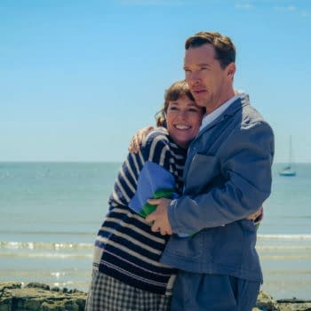 A scene from 'The Roses' featuring actors Olivia Colman and Benedict Cumberbatch embracing against a beach backdrop with gentle waves and a clear blue sky.
