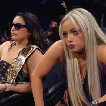 WWE Women's Tag Champions Raquel Rodriguez and Liv Morgan sit attentively during a tag team gauntlet match on WWE SmackDown. Raquel wears sunglasses and a championship belt, while Liv appears focused and engaged.