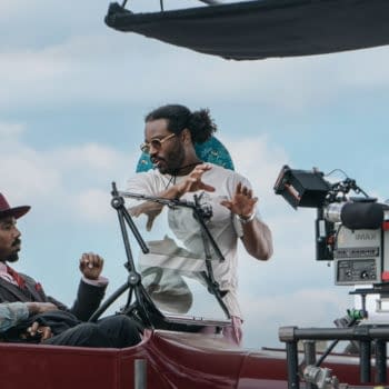 A scene from a film set featuring Ryan Coogler engaging with two actors seated in a vintage car, while an IMAX camera is positioned nearby. The sky is partly cloudy, and the atmosphere suggests a behind-the-scenes look at filmmaking.