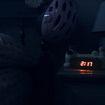 A dimly lit bedroom at night featuring a bedside table with a digital clock displaying 2:17, various small plush toys, a lamp with a horse figurine, and a pink bicycle helmet hanging nearby.