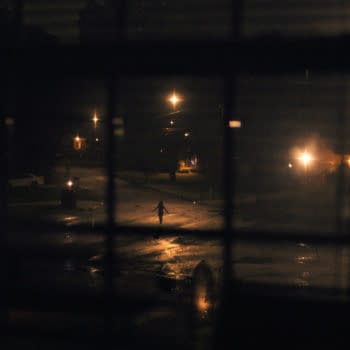 A dark scene viewed through a window, featuring a figure walking down a rain-soaked street illuminated by streetlights, creating a dramatic contrast against the night.