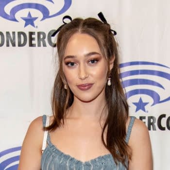 A person with long brown hair styled with black ribbons is standing in front of a WonderCon backdrop. They are wearing a light blue, ruffled top and earrings.