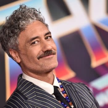 A celebrity, Taika Waititi, smiles while wearing a striped suit and colorful beads on a red carpet at a movie premiere, against a vibrant backdrop.