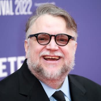 A portrait of Guillermo del Toro, the acclaimed Mexican filmmaker, smiling widely while wearing black glasses and a dark suit. The background features a purple banner for a 2022 festival.