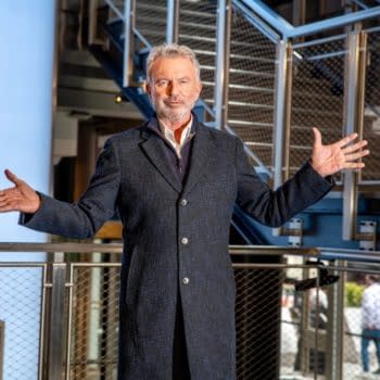 Actor Sam Neill stands in the Empire State Building with arms open, showcasing the iconic architecture around him.