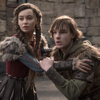A scene from 'How To Train Your Dragon' featuring two characters, a young woman with braided hair in a red and grey costume, and a young man with tousled hair in a green hoodie and fur-trimmed clothing, looking distressed while holding onto each other in a medieval setting.