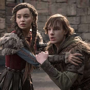 A scene from 'How To Train Your Dragon' featuring two characters, a young woman with braided hair in a red and grey costume, and a young man with tousled hair in a green hoodie and fur-trimmed clothing, looking distressed while holding onto each other in a medieval setting.