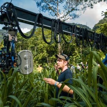 On the set of 'Jurassic World Rebirth,' director Gareth Edwards appears engaged while surrounded by tall green grass and a crew. A camera on a crane is positioned above, capturing the action in a lush outdoor environment.