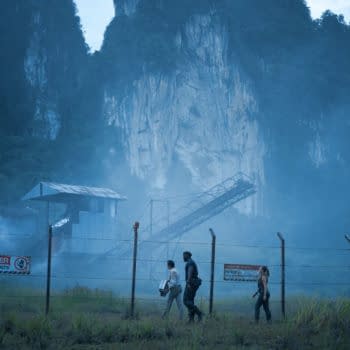 A scene from 'Jurassic World Rebirth' shows four characters walking along a foggy, overgrown area near a chain-link fence. In the background, a dilapidated building and rocky cliffs are visible, creating an eerie atmosphere.