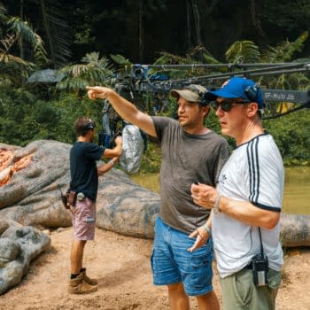 On the set of 'Jurassic World Rebirth,' director Gareth Edwards points while discussing a scene, flanked by crew members and a large, dinosaur-like prop in a lush, green environment.