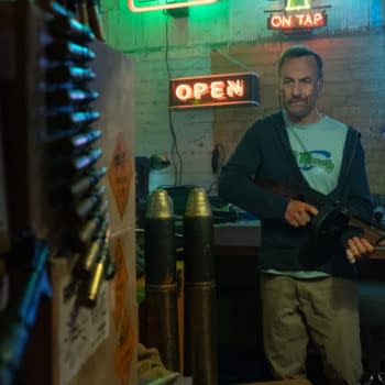 Bob Odenkirk portrays Hutch Mansell holding a gun in a dimly lit room featuring neon signs, surrounded by tools and military artifacts, in a scene from the film Nobody 2.