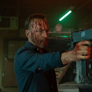 A man with a bloodied face and hands aims a gun in a dimly lit, industrial setting. The atmosphere is tense, highlighted by a green neon light.