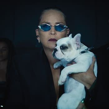 A woman in a black suit holds a small white French bulldog, wearing sunglasses. She is flanked by two figures in dark clothing, against a dimly lit background.