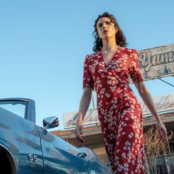 Margaret Qualley walks confidently past a vintage blue car, wearing a red floral dress, with a dilapidated shop sign in the background that reads "Yums Bakery." This scene is from Ethan Coen's film "HONEY DON'T!".