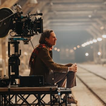 Wes Anderson sits on set with a film camera on a tripod in the foreground, surrounded by a dimly lit railway line that features overhead lights, during the production of 'The Phoenician Scheme.'
