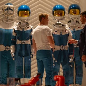 A scene from 'The Fantastic Four: First Steps' featuring two characters, Joseph Quinn as Johnny Storm and Pedro Pascal as Reed Richards, interacting in front of futuristic blue astronaut suits, emphasizing the Sci-Fi theme. The suits prominently display the number '4' symbol.