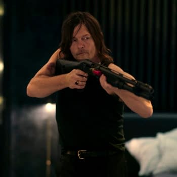 A man with long hair is aiming a shotgun, wearing a sleeveless black shirt and looking intensely focused. He stands in a dimly lit room with a bed in the background, suggesting a tense or dramatic scene.