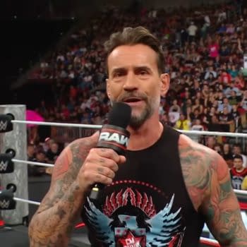 A wrestler with a tattooed arm speaks into a microphone while standing in a wrestling ring. The audience is visible in the background, showing excitement during the live event.