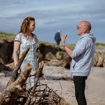 A behind-the-scenes moment on a seaside set, featuring a woman in a floral dress engaging in conversation with a man wearing a gray hoodie. A crew member is seen in the background, alongside driftwood scattered on the beach.