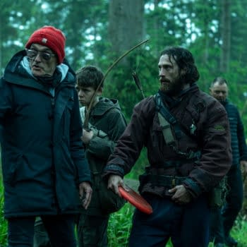 A group of four people in a forest, with one wearing a red beanie and gesturing, while another holds an arrow and bow. The scene conveys a sense of urgency and focus, suitable for a narrative about survival or adventure.