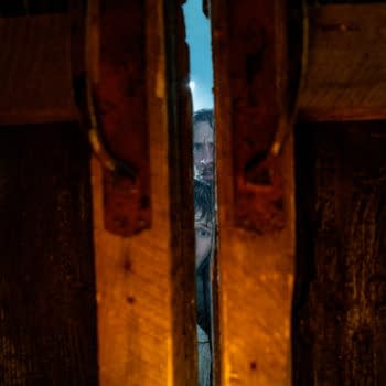 A cinematic scene viewed through a crack in wooden planks, revealing two faces with a tense expression. The atmosphere is moody and suspenseful, suggesting a moment of anticipation.