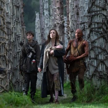 A scene from the post '28 Years Later:' featuring a boy holding hands with a woman who carries a baby, accompanied by a muscular man, all walking through a dense forest of tall, unique trees with bark resembling bones.