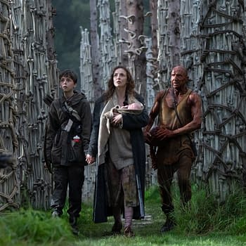 A scene from the post '28 Years Later:' featuring a boy holding hands with a woman who carries a baby, accompanied by a muscular man, all walking through a dense forest of tall, unique trees with bark resembling bones.