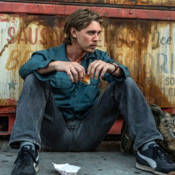 A young man in a teal shirt sits on the ground against a rusty backdrop, appearing contemplative while holding a piece of food. Nearby, a cat peeks out from a bag, showing a defensive posture.