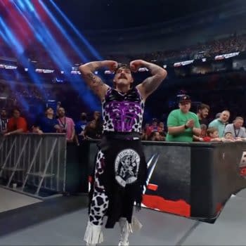 Dominik Mysterio flexes his muscles confidently after executing a successful maneuver during a WWE Backlash event, surrounded by cheering fans and colorful stage lights.