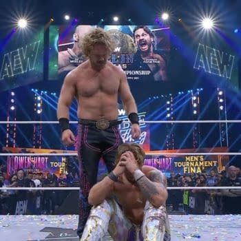 Hangman Adam Page stands triumphantly over a kneeling Will Ospreay in a wrestling ring, celebrating his victory. Confetti is scattered on the canvas, with colorful lights illuminating the arena in the background.