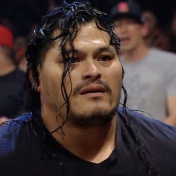 A man with long, wet hair has a shocked expression, surrounded by a lively crowd during a wrestling event. He appears to be making his debut as a wrestler.