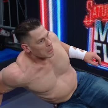 John Cena, with a shocked expression, is seated on the floor in front of a backdrop featuring the WWE Saturday Night's Main Event logo. He is shirtless and wearing denim shorts, with visible muscles and a bandage on his arm.