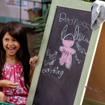 Lilo, played by Maia Kealoha, is joyfully laughing next to a chalkboard with a drawing of Stitch and text that reads 'Don't break everything' and 'Lend a helping hand.' This scene is from Disney’s live-action Lilo & Stitch.