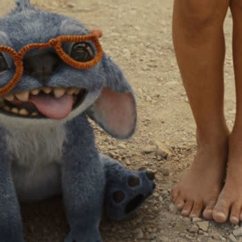 Stitch, a blue alien character from Disney's live-action 'Lilo & Stitch,' is sitting on the ground, wearing orange glasses and playfully sticking out his tongue alongside a child's bare feet.