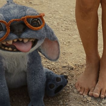 Stitch, a blue alien character from Disney's live-action 'Lilo & Stitch,' is sitting on the ground, wearing orange glasses and playfully sticking out his tongue alongside a child's bare feet.