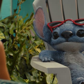 In Disney's live-action Lilo & Stitch, Stitch, the blue alien character, wears red sunglasses and relaxes in a white lawn chair, facing Lilo, who is partially visible in the background surrounded by greenery.