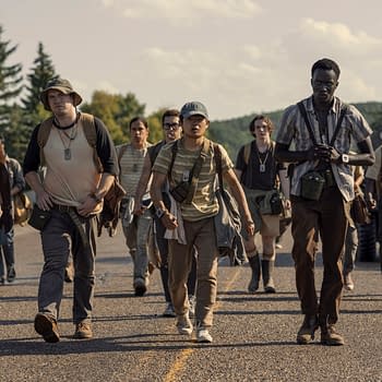 A group of young men, including characters portrayed by David Jonsson, Cooper Hoffman, and Jordan Gonzalez, are walking together along a road, dressed in casual outdoor clothing with backpacks and gear. The scene reflects a tense atmosphere as they embark on a significant journey in The Long Walk.