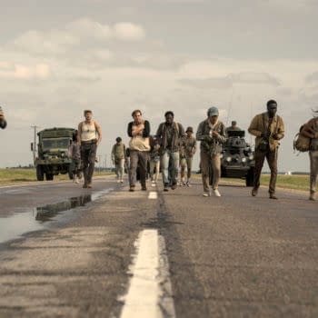 A scene from 'The Long Walk' featuring a group of young men walking on a highway, with military vehicles in the background. The characters appear determined as they traverse the road under a cloudy sky.