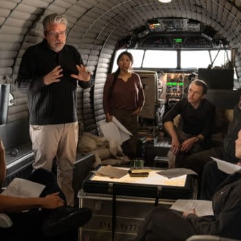 A scene from 'Mission: Impossible - The Final Reckoning' showing a group of six involved in a discussion on a set designed like an aircraft interior. The director is gesturing while actors focus on the conversation, with scripts and a table of documents visible.