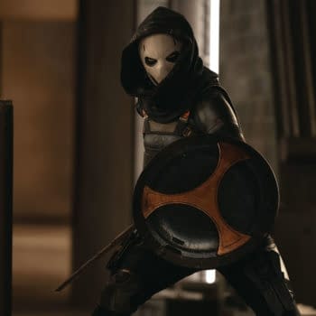 A character dressed as Taskmaster, portrayed by Olga Kurylenko, wears a hood and a mask while holding a shield, in a scene from Marvel Studios' THUNDERBOLTS*. The background features industrial elements.