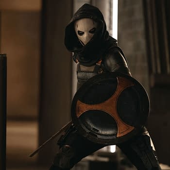 A character dressed as Taskmaster, portrayed by Olga Kurylenko, wears a hood and a mask while holding a shield, in a scene from Marvel Studios' THUNDERBOLTS*. The background features industrial elements.