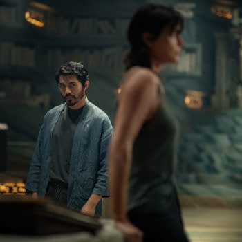 A scene from 'The Old Guard 2' featuring Henry Golding as Tuah in a blue robe, looking intently at Charlize Theron as Andy, who is in the foreground. The setting is dimly lit with shelves of books in the background.