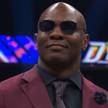 A confident Shelton Benjamin wearing sunglasses and a stylish dark suit with a patterned tie, standing on the set of AEW Dynamite. The background features the AEW Dynamite logo in vibrant colors.