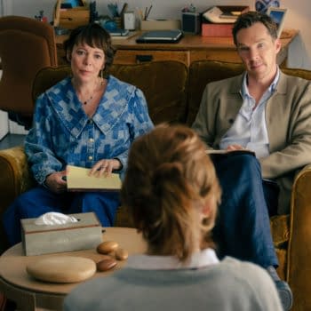 A scene from 'The Roses' featuring Olivia Colman and Benedict Cumberbatch sitting on a couch, engaged in conversation with a person out of frame. The setting includes a cozy living room with a coffee table displaying a tissue box and smooth stones.