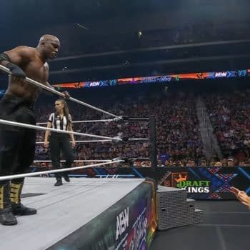A tense moment at AEW Double or Nothing, featuring a wrestler in black attire standing next to the ropes, while another man in a blue suit gestures toward him. The ring is surrounded by a cheering crowd and a referee watches attentively.