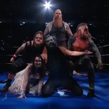 A group of wrestlers, dressed in spooky and dark attire, poses dramatically in a wrestling ring under bright lights, with a dark audience in the background.